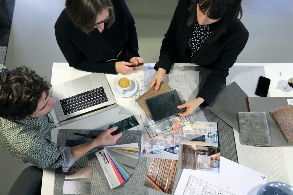 Bovenaanzicht van drie personen die samen materiaalstalen en ontwerpen bespreken aan een tafel, met laptop, smartphone en kleurstalen erbij.