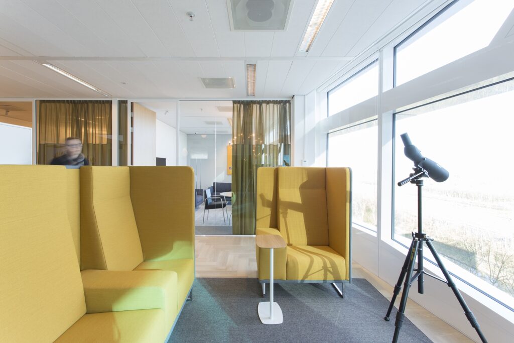 Een lichte kantoorruimte met twee hoge, gele loungefauteuils tegenover elkaar en een klein wit tafeltje ertussen. Rechts staat een telescoop bij het raam. Op de achtergrond is een vergaderruimte zichtbaar achter groene transparante gordijnen.