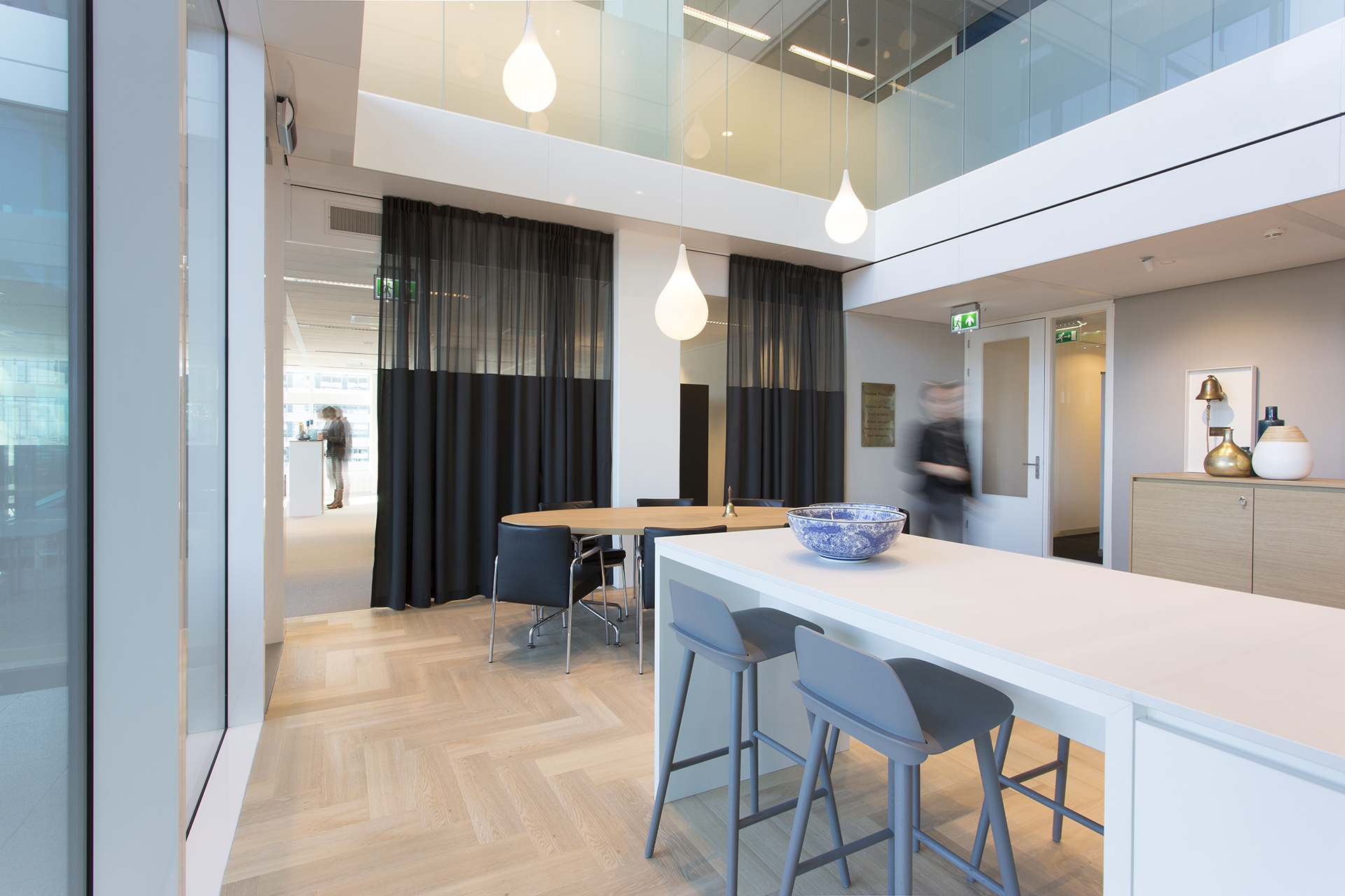 Een moderne kantoorruimte met een hoge vide en lichte houten visgraatvloer. Op de voorgrond staat een wit kookeiland met barkrukken en een schaal erop. Achterin bevindt zich een vergaderruimte met een tafel en stoelen, deels afgescheiden door donkere gord