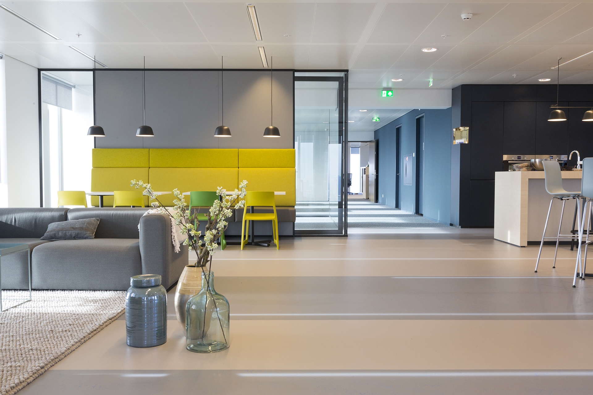 Een moderne kantoorruimte met een zithoek en een gele wandbank onder drie hanglampen. Op de voorgrond staan glazen vazen met bloemen op een tafel. Rechts loopt een gang met blauwe wanden en meerdere deuren. De ruimte heeft een lichte, strakke afwerking en