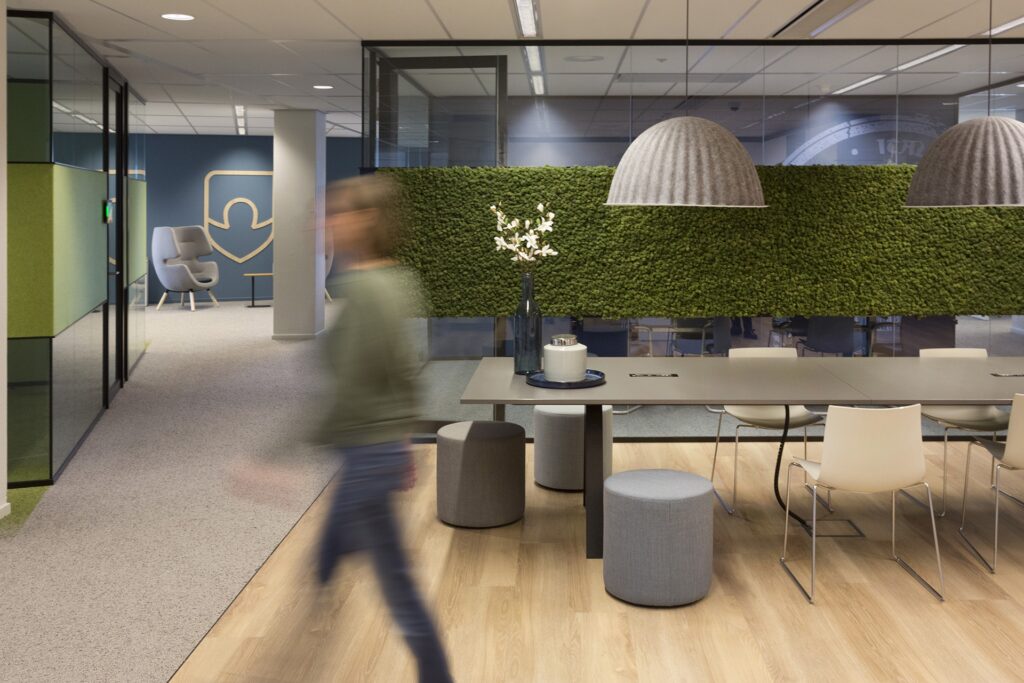 Een moderne kantoorruimte met een lange tafel, poefjes en stoelen op een lichte houten vloer. Aan de achterwand bevindt zich een grote groene plantenwand. Een persoon loopt onscherp door het beeld. Boven de tafel hangt een grote ronde hanglamp.