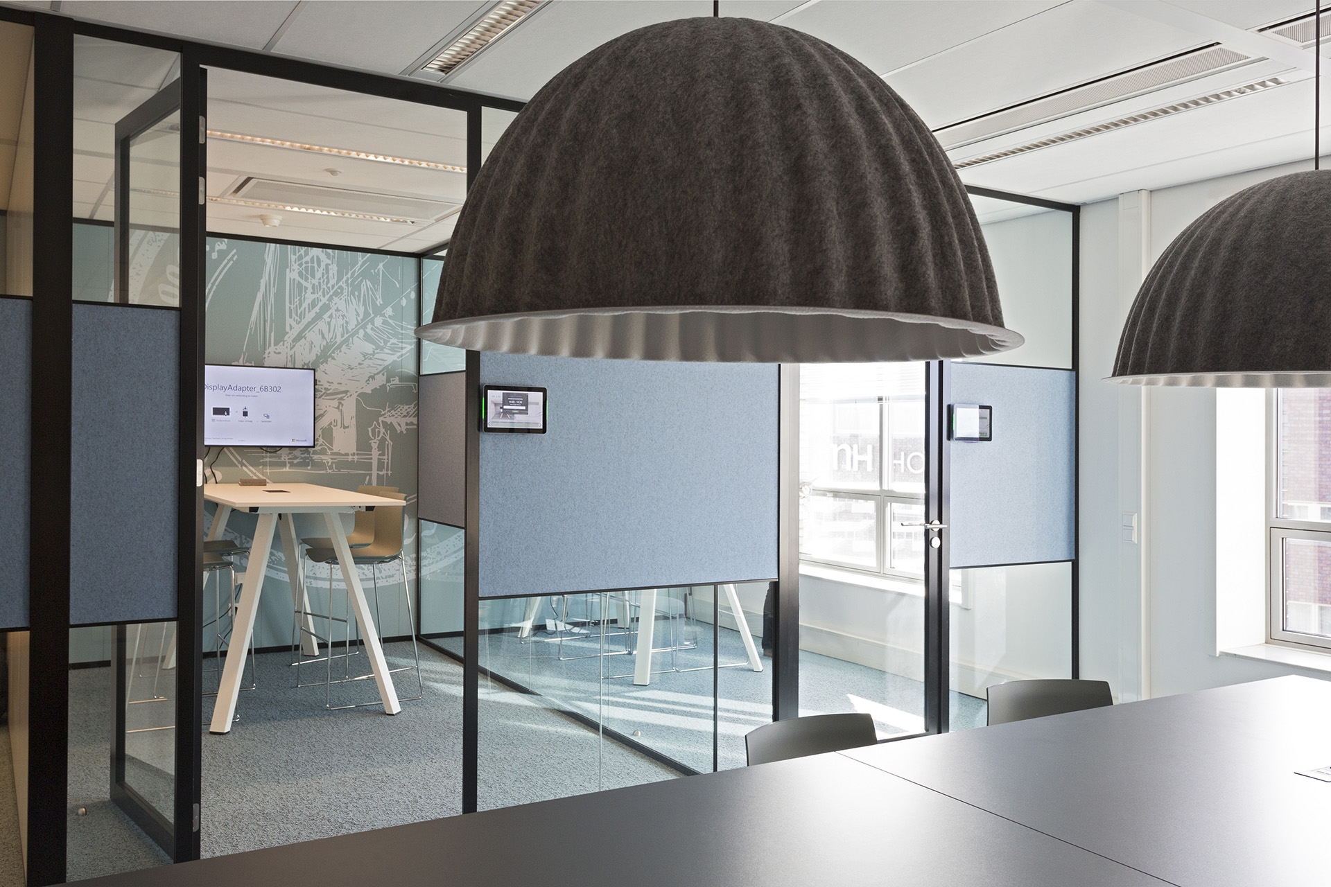 Een moderne kantoorruimte met glazen vergaderruimtes en zwarte frames. Op de voorgrond hangen twee grote donkere hanglampen boven een tafel. Achter de glazen wanden zijn werkplekken zichtbaar met hoge tafels en blauwe panelen. De ruimte heeft een strakke