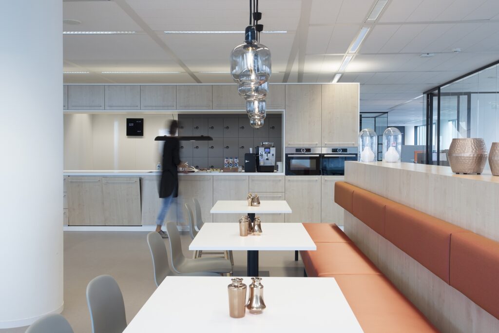 Een moderne kantoorpantry met lichte houten kasten en ingebouwde apparatuur. Op de voorgrond staan witte tafels met een vaste zitbank in oranje tinten. Een persoon loopt onscherp langs het aanrecht. Boven de tafels hangen glazen designlampen.