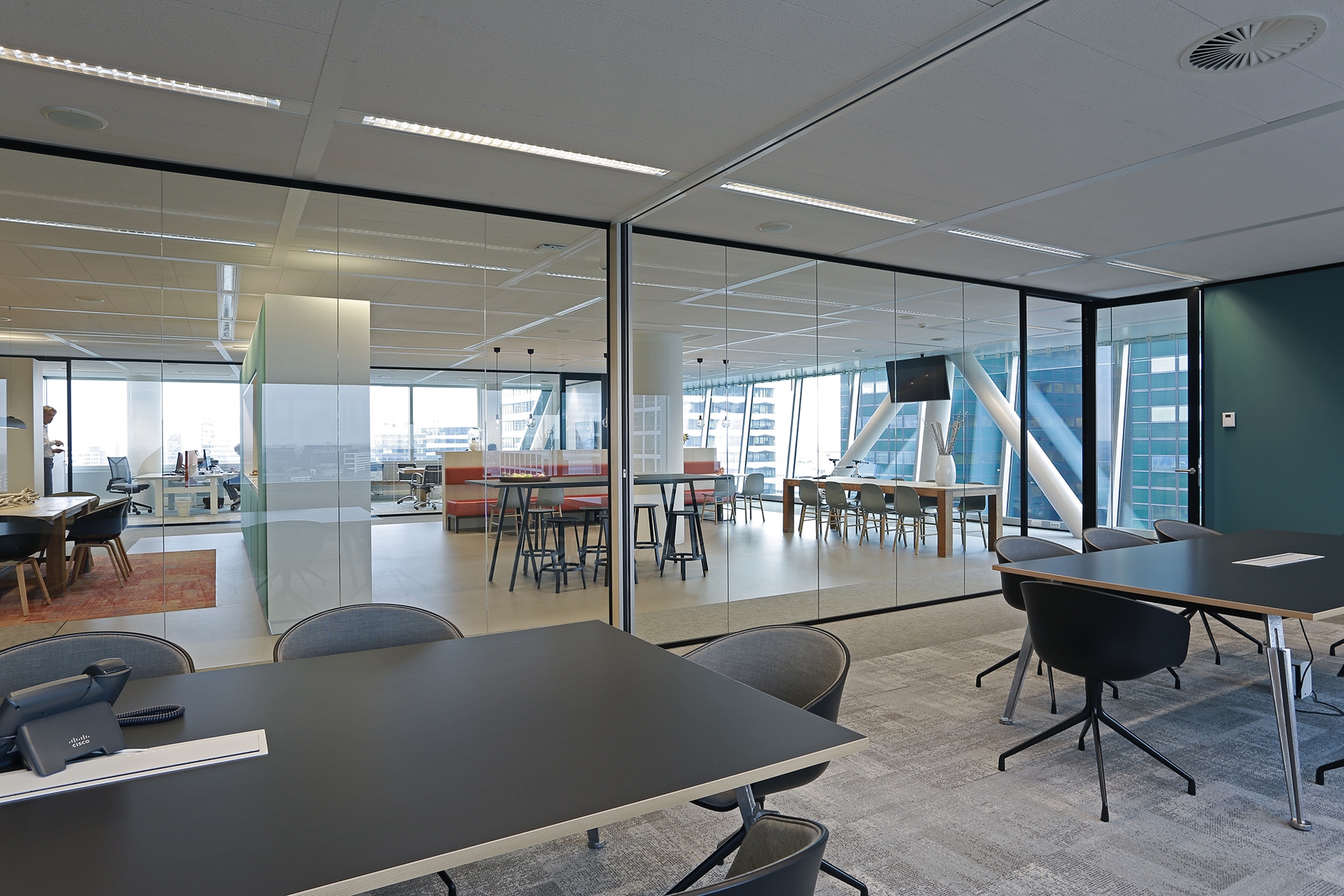 Een moderne kantoorruimte met zwarte tafels en stoelen op de voorgrond. Achter glazen wanden is een open werk- en ontmoetingsruimte zichtbaar met hoge tafels en grote ramen. De ruimte heeft een strakke afwerking en veel daglicht.