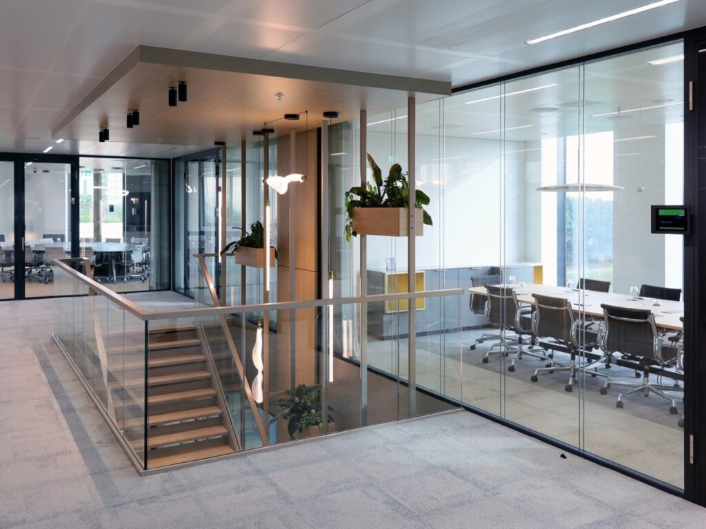 Een moderne kantoorruimte met een glazen wand en vergaderruimte, voorzien van bureaustoelen en een lange tafel.