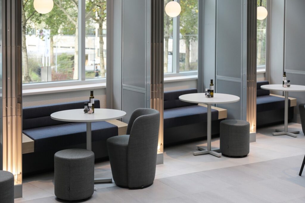 Moderne loungehoek met ronde tafels, gestoffeerde zitplaatsen en grote ramen in een rustige kantooromgeving.