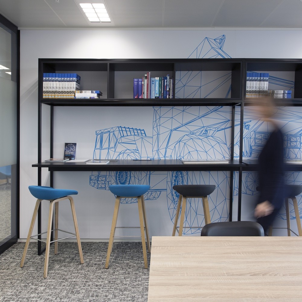 Een moderne kantoorruimte met een witte muur waarop een blauw geometrisch ontwerp van een vrachtwagen is afgebeeld, drie hoge krukken en een voorbijlopende persoon.