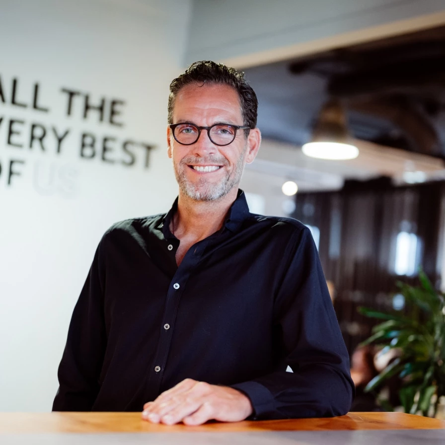 A man in a black shirt with glasses is smiling while leaning on a desk in an office setting.