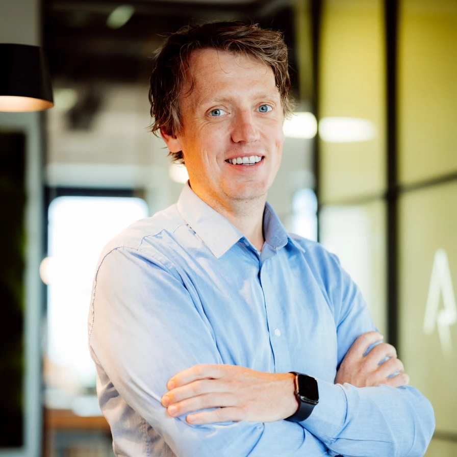 Een man in een blauwe overhemd poseert met gekruiste armen in een moderne kantooromgeving.