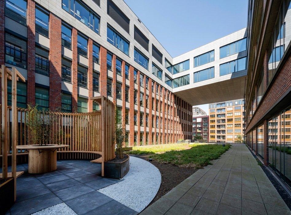 Een moderne binnentuin omringd door een kantoorgebouw met een gevel van baksteen en grote ramen. In de tuin staan planten en een houten bank met een gebogen rugleuning. De gebouwen vormen een open binnenplaats met een doorgang naar een ander complex.