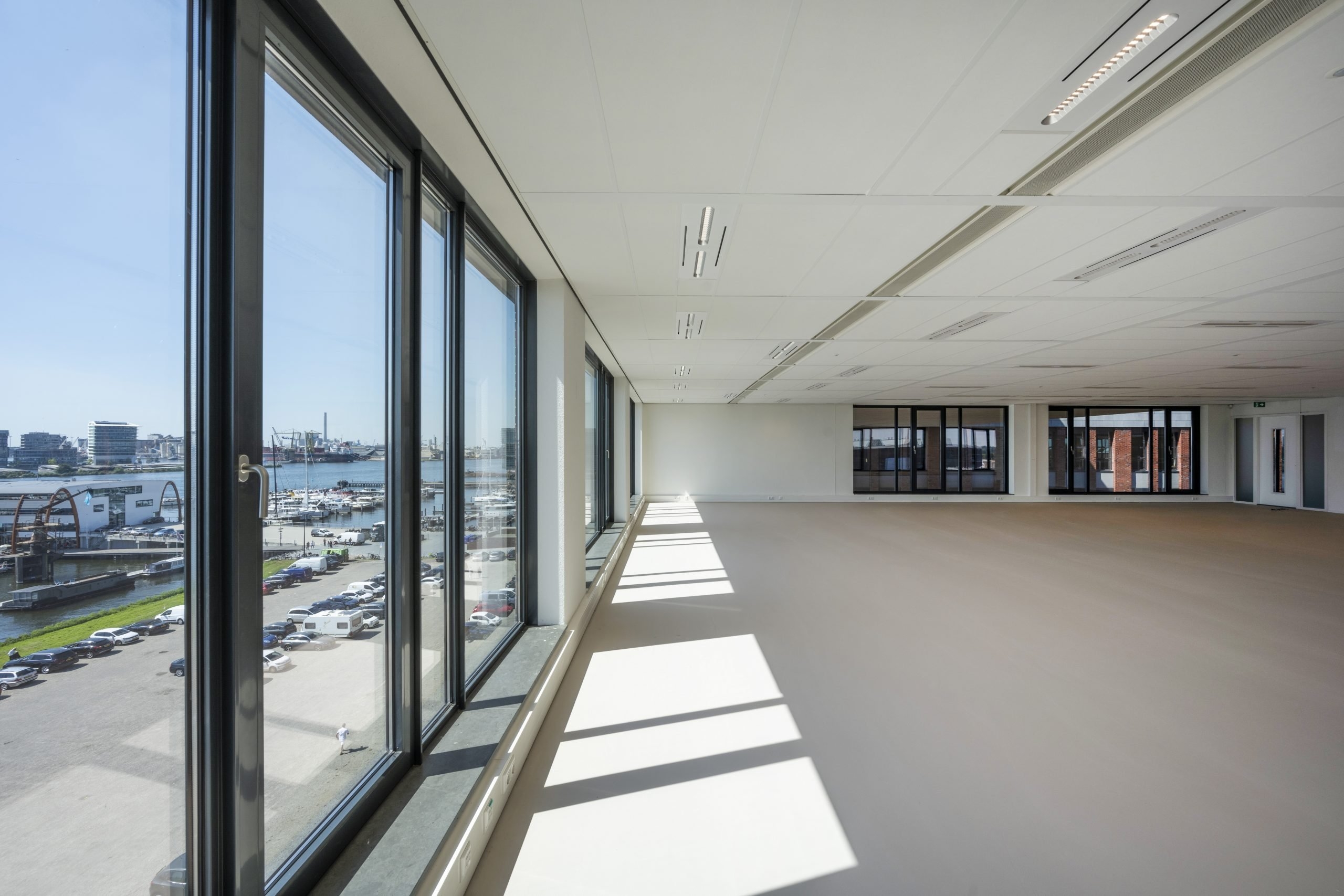 Een grote, lege kantoorruimte met vloer-tot-plafondramen langs één zijde. Zonlicht valt naar binnen en werpt lange schaduwen op de vloer. Door de ramen is uitzicht op een parkeerplaats en omliggende gebouwen zichtbaar. De ruimte heeft een strak plafond me
