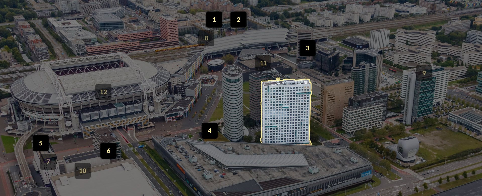 Een luchtfoto van een stedelijk gebied met meerdere grote gebouwen en wegen. Eén hoog, rechthoekig gebouw is met een lichte omlijning gemarkeerd. Op verschillende locaties in de afbeelding staan zwarte labels met witte cijfers die specifieke gebouwen aand