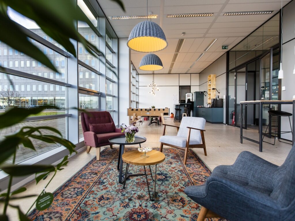 Twee collega’s zitten aan een ronde houten tafel in een moderne vergaderruimte met glazen wanden. Eén persoon maakt aantekeningen in een notitieboek terwijl de ander meedenkt, wat een rustige en samenwerkende werksfeer weergeeft.