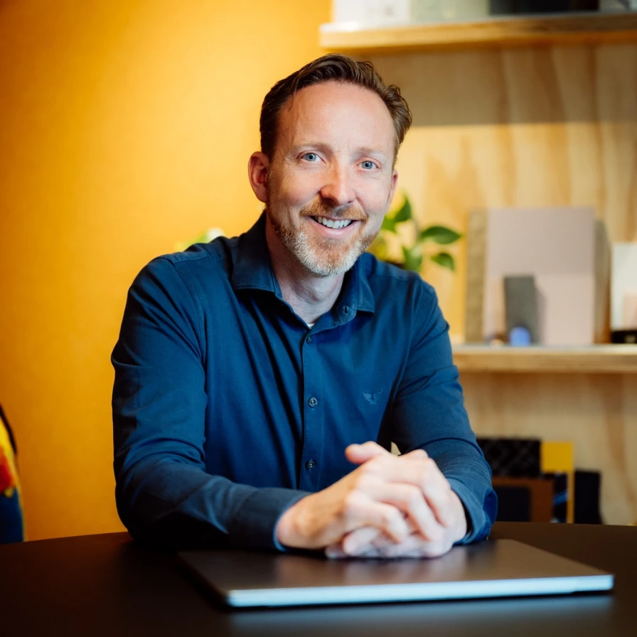 A smiling man in a blue shirt sits at a table with a closed laptop.