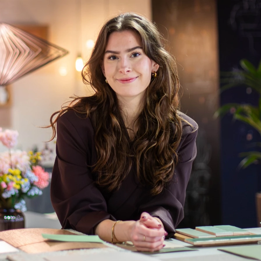 A woman with long brown hair smiles while leaning on a table in a warmly lit room decorated with flowers.