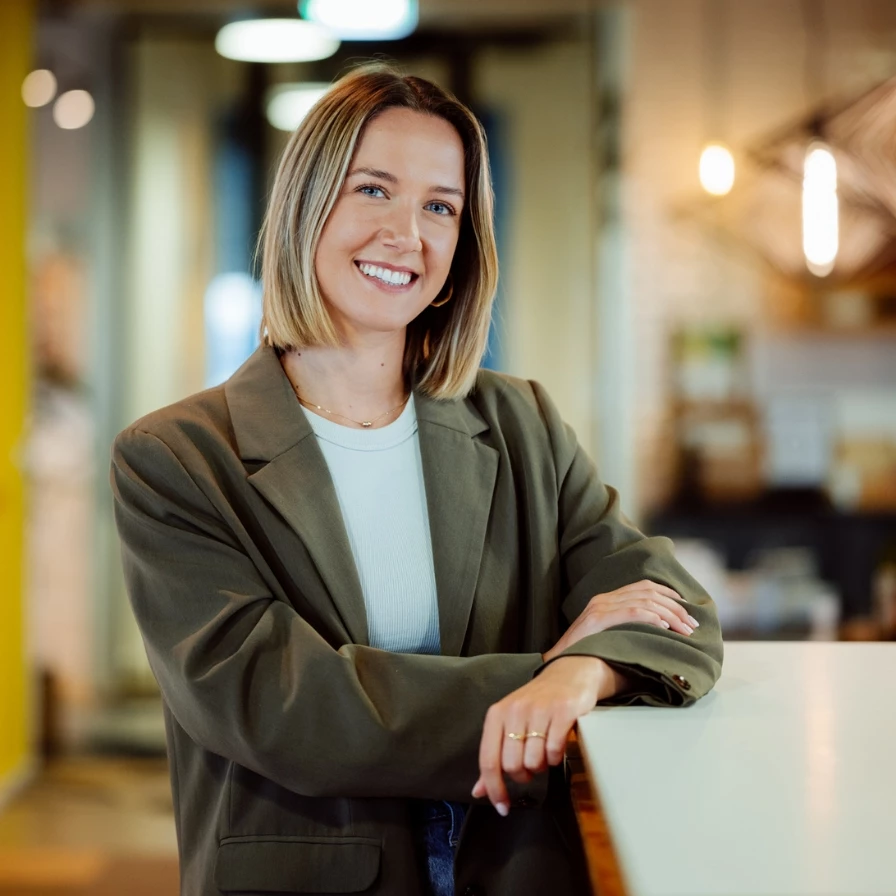 Vrouw in een groene blazer glimlacht en leunt tegen een balie in een modern interieur.