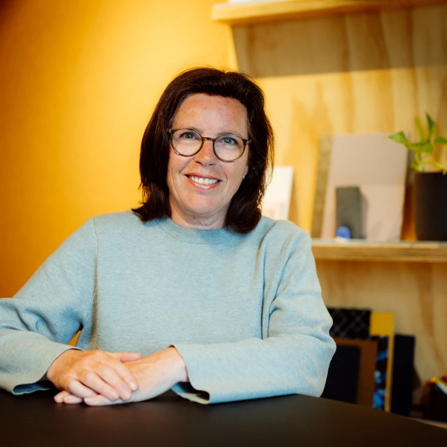 Vrouw met bril en glimlach zittend aan een tafel met een houten achtergrond.