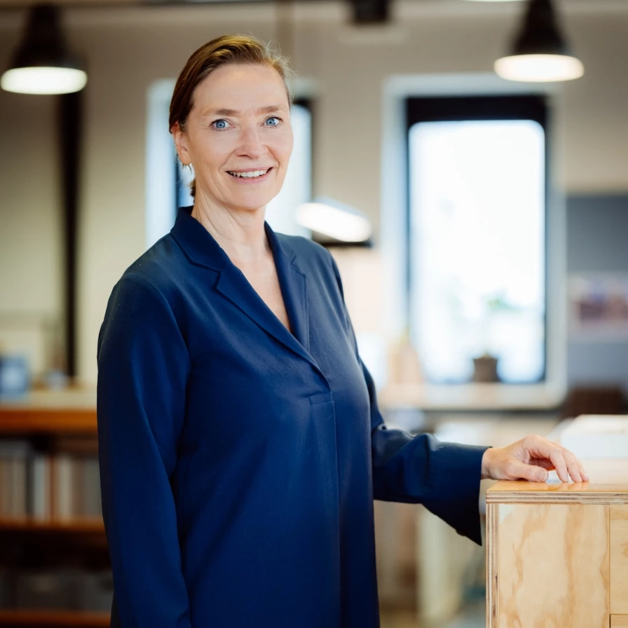 Een lachende vrouw in een donkerblauw kledingstuk poseert in een moderne kantooromgeving.