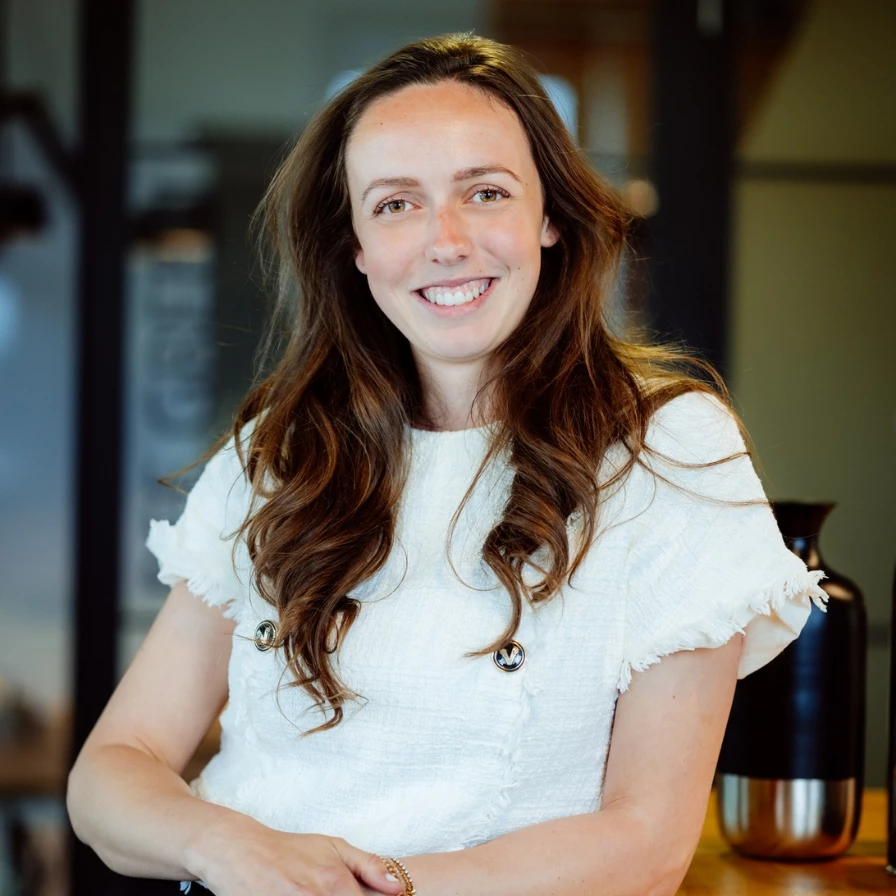 A woman with long brown hair wearing a white top smiles while leaning against a table indoors.
