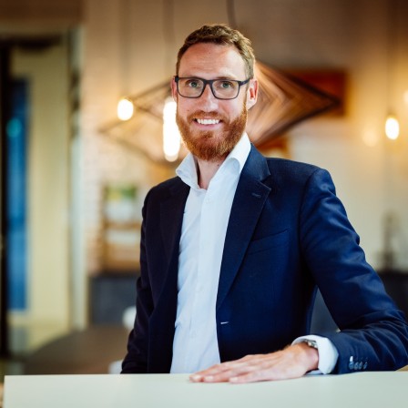 Een lachende man met een bril en baard in een zakelijke outfit staat bij een tafel in een modern ingerichte ruimte.