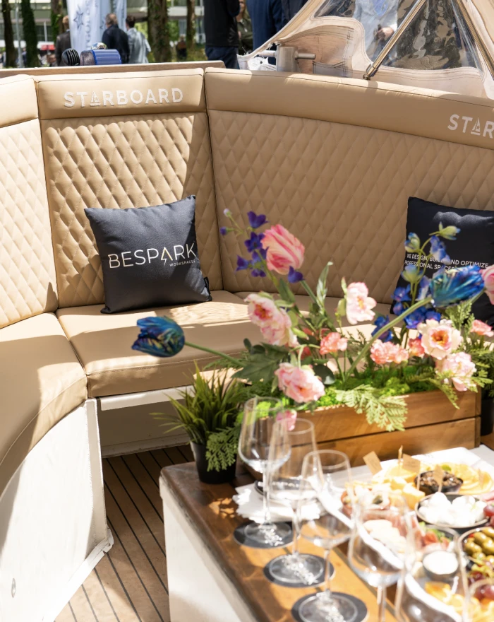 Luxe bootinterieur met beige gestoffeerde stoelen, blauwe kussens met het logo van "BESPAK," en een houten tafel gedecoreerd met bloemen en glazen.