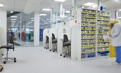 Een modern laboratorium met werkstations en een rek vol potjes, waar een persoon in een witte jas en blauwe broek bezig is.