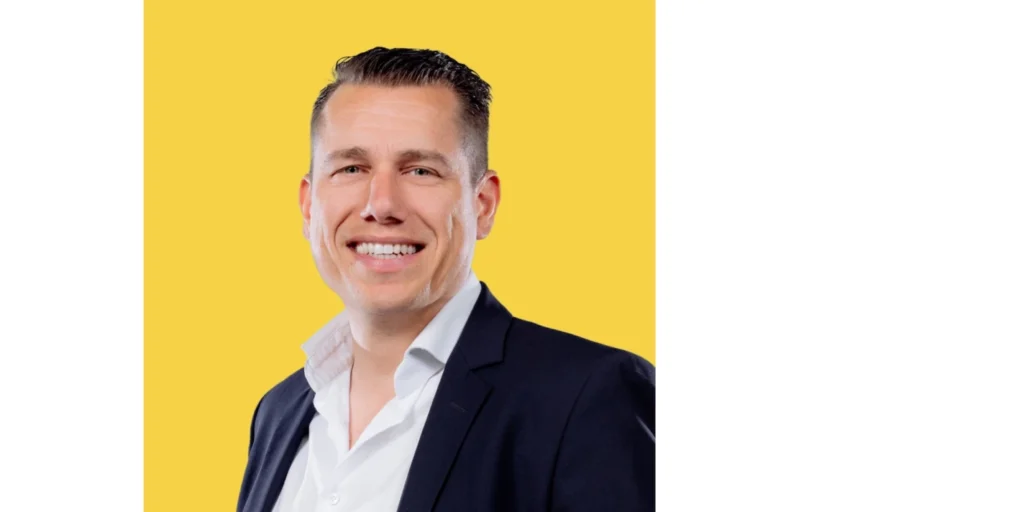 Man in een donker pak met een witte overhemd, glimlachend voor een gele achtergrond.