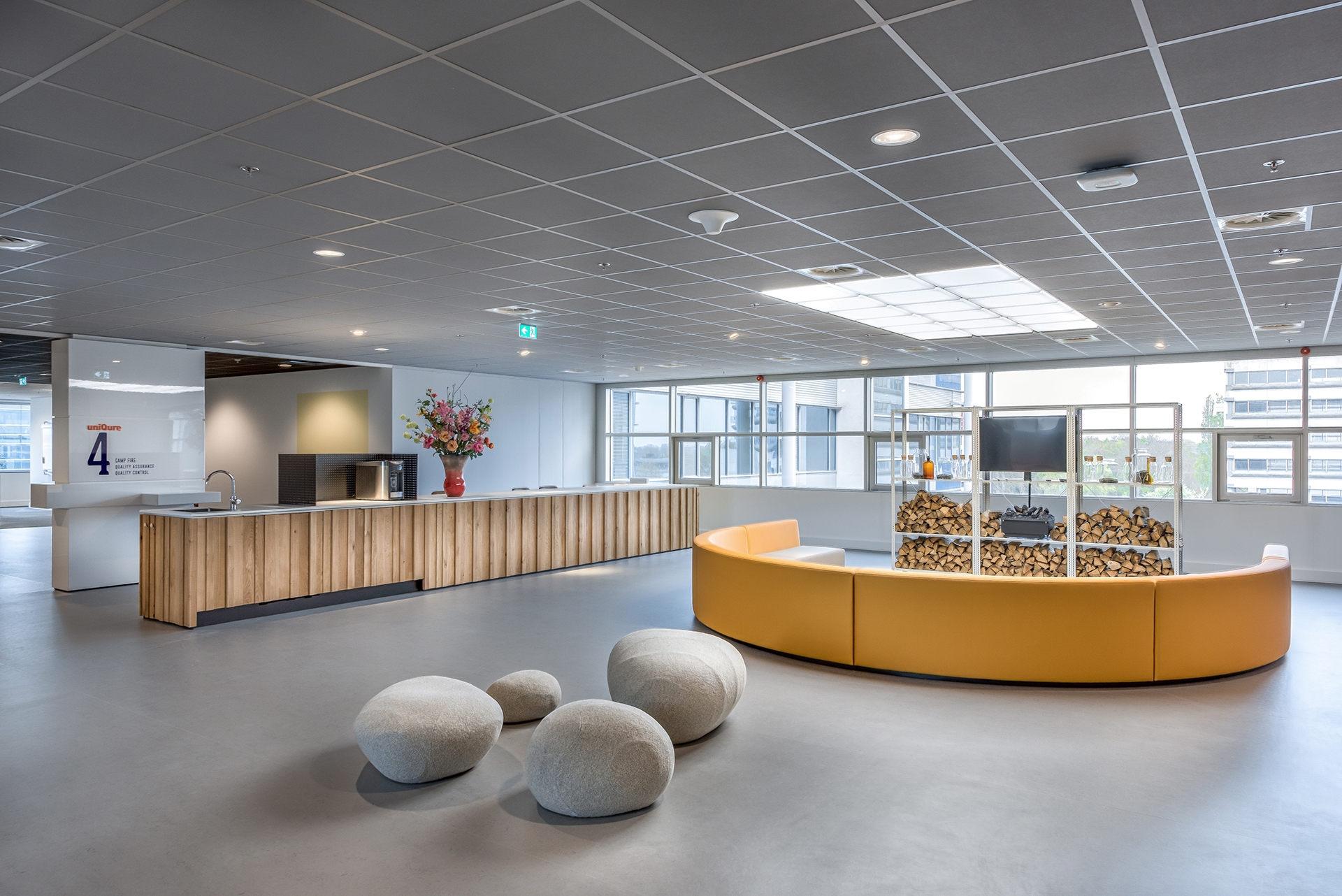 Een moderne ontvangsthal met een lange houten receptiebalie aan de linkerzijde. In het midden staat een ronde gele zitbank met een glazen vitrine erop. Op de voorgrond liggen enkele ronde zitpoefjes. Grote ramen zorgen voor veel natuurlijk licht in de rui