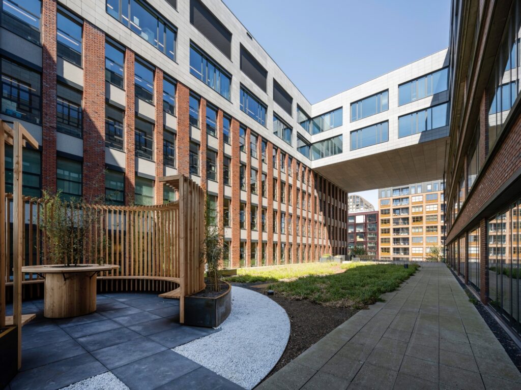Binnenplaats van een modern gebouw met gevel van baksteen en glas op de NDSM-werf, met een houten zithoek en groene beplanting.
